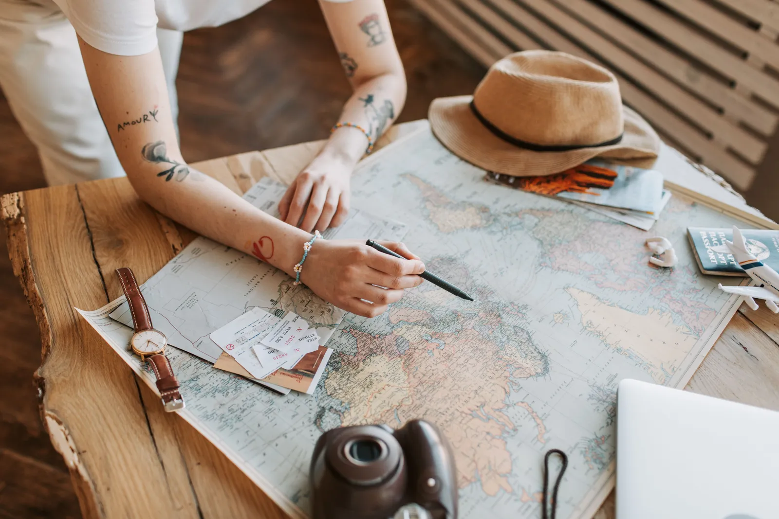 Tattooed woman planning a trip with a world map, passports, and travel essentials on a wooden table